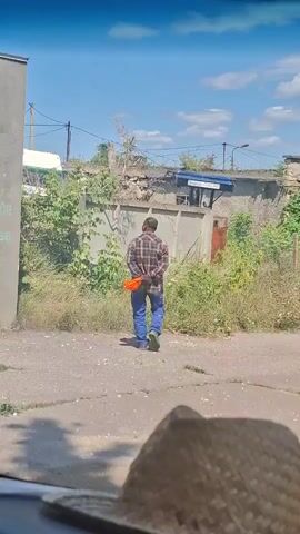 A man wearing a plaid shirt and blue jeans is standing in an outdoor area. He is holding an orange object behind his back with his hands. The area appears to be a street or an alleyway with a concrete surface. There is overgrown vegetation and a wall in the background. A straw hat is visible in the foreground, partially obscuring the lower portion of the image. The sky is blue with some clouds.