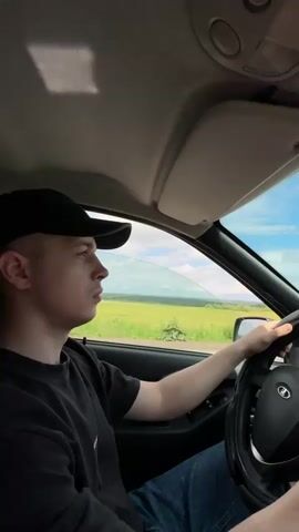 A young man wearing a black baseball cap and a black t-shirt drives a car. The sun shines through the windshield, casting a shadow on the dashboard. Outside, a field of green crops stretches to the horizon under a blue sky with scattered clouds. The man's hands are on the steering wheel, and he appears to be focused on the road. The interior of the car is visible, including the dashboard, steering wheel, and part of the passenger seat.