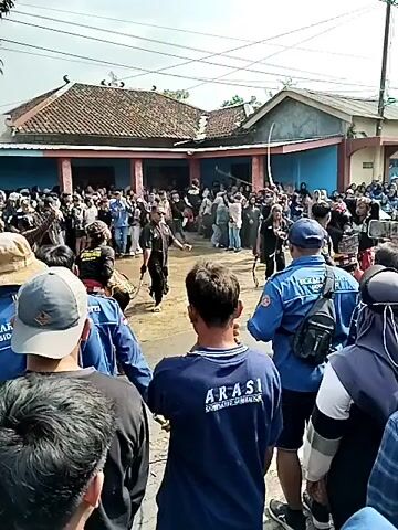 A crowd of people is gathered on a street in front of a building with a red tile roof. Some people in the crowd are wearing blue shirts with "ARASI JAWA TIMUR" written on the back. In the center of the street, several individuals are engaged in what appears to be a performance or ritual. One person is wearing traditional clothing, including a patterned sarong and a black shirt, and is holding a whip. Another person in a black shirt with "PESILAT" on the back is also present. The performance involves movement and interaction among the participants. Some spectators are holding up phones to record the event. The environment appears to be outdoors with a clear sky. The scene suggests a public gathering or cultural event taking place in a village or small town.