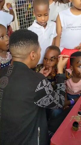 A man is drawing a triangle on a child's forehead with a thin brush. The child is sitting at a red table, and two other children are sitting beside them. The child on the right has whiskers drawn on their face. Other children are standing behind the group, watching the activity. Jars of paint and glitter are on the table. The man is wearing a black jacket with white designs. The music in the background is lively.