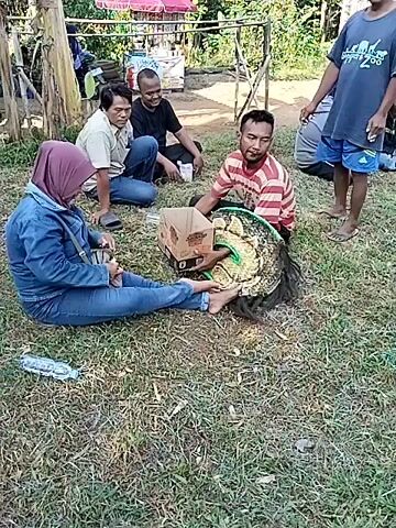 The video shows a group of people gathered on a grassy area outdoors. Some are sitting on the grass, while others are standing. A man in a striped shirt holds a cardboard box and a Barongan mask. Another person hands the man something while sitting on the ground. A small child stands nearby, looking at the man with the mask. The background includes trees, a building, and various other objects.