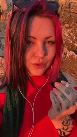 A woman with pink and dark hair stands near a rocky area and exhales vapor. She wears a red shirt, green scarf, and sunglasses. She has white earphones in. The scene is illuminated by what appears to be late afternoon sunlight.