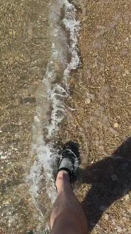 The video shows a person standing in shallow water on a beach. The person is wearing dark-colored sandals and the camera is positioned looking down at their feet and the water. The water is clear and shallow, revealing the sand and small pebbles on the beach floor. The waves are gently lapping at the shore. The person is standing still, allowing the water to wash over their feet. Their shadow is cast onto the wet sand. The scene suggests a calm, sunny day at the beach.