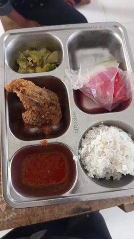 A top-down shot shows a stainless steel cafeteria tray divided into five compartments, sitting on a brown wooden surface. The compartments contain a small portion of green vegetables (likely broccoli and corn), a piece of fried chicken, a red sauce, a small plastic bag of watermelon slices, and a portion of white rice. The tray has "SUS304" stamped into the metal near the rice compartment. In the background, two people are partially visible, both wearing dark pants and appearing to be seated. There is ambient background noise.