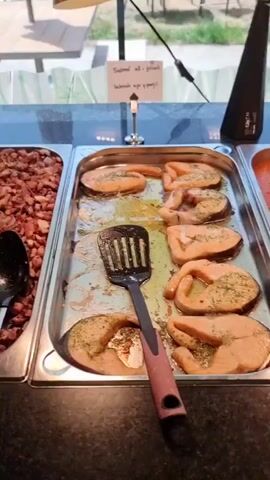 The video shows a buffet line with various food items. On the left, a silver tray contains several slices of salmon with herbs. Next to it, a larger silver tray holds pieces of fish in a reddish sauce. To the right of that, another silver tray displays cooked fish pieces, and next to it, a tray is filled with red peppers in sauce. Further to the right, a glass bowl contains noodles, and next to that, a tray holds what appears to be a baked dish. In the background, there are more empty buffet trays and a window looking out onto a grassy area.