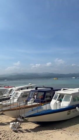 The video shows a row of speedboats docked on a sandy beach. The boats are white with blue trim and have stairs leading up to them. There are people on the beach and on some of the boats, seemingly preparing for or disembarking from a trip. In the background, there is a clear blue sky with scattered clouds, calm turquoise water, and a distant shoreline with mountains. The overall scene depicts a sunny day at a tropical beach resort.