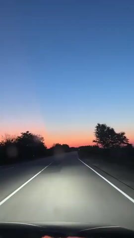The video shows a first-person view from inside a car driving on a paved road at dusk. The sky is a gradient of deep blue at the top, transitioning to orange and pink near the horizon, indicating the setting sun. The road has white dividing lines, and the car's headlights illuminate the path ahead. Silhouettes of trees and bushes line both sides of the road. The camera is focused forward, capturing the ongoing journey.
