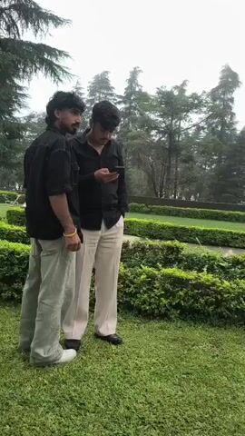 The video shows a group of people in an outdoor setting. In the foreground, two young men in black shirts and light-colored pants are standing, with one of them looking at his phone. To the right, another young man in a white shirt and black pants is standing in a garden area, touching his hair and looking around. Further in the background, there are trees and a building. The environment appears to be a park or garden with manicured hedges and green grass. The sky is overcast.