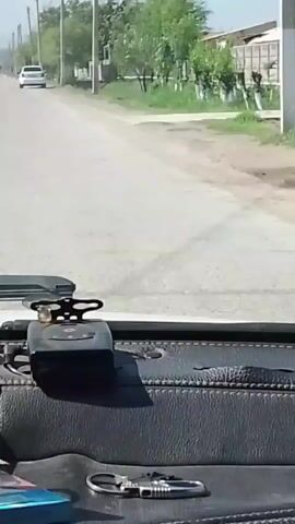 The video shows a point-of-view shot from inside a vehicle driving on a paved road. The road is straight with a white car in front. There are trees and utility poles along the side of the road. The dashboard of the vehicle is visible with a cigarette pack, keys, and other small items on it. The weather appears to be sunny and clear.  A truck is seen in the distance. There is a bumpy ride.