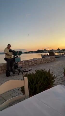 A man stands playing a keyboard on a wheeled cart on a paved walkway next to a stone wall overlooking a body of water. A sunset is visible in the background with a few boats docked in the water. Green plants are in the foreground. A white chair is partially visible in the bottom corner of the frame.