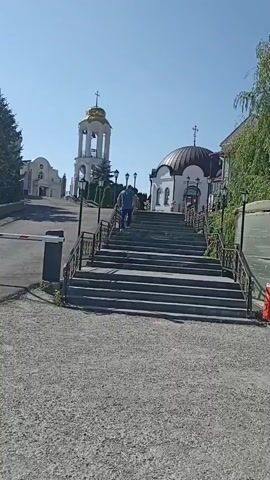 A low-angle, shaky, full-shot video shows a person walking up a wide, stone staircase towards a church. The church has a dome with a cross on top. To the left of the church is a tall, white bell tower with a gold dome. There are trees and bushes around the church and bell tower. A person in a blue shirt and black pants is walking up the stairs. There is a red trash can next to a structure with a sign written in Cyrillic. The sky is clear and blue.