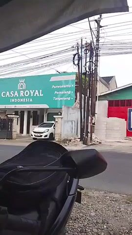 The video opens with a view from behind a motorcycle, focusing on its handlebars and a black helmet hanging from the left handlebar. The background shows a street with a building labeled "CASA ROYAL INDONESIA" on a large green sign. A white car is parked in front of the building. Electrical poles and wires are visible. A red van drives past on the street. Another motorcycle with two riders passes by. The perspective shifts slightly, showing more of the street and surrounding buildings. The motorcycle in the foreground has "HONDA" visible on its body. The scene appears to be an outdoor environment on a cloudy day.
