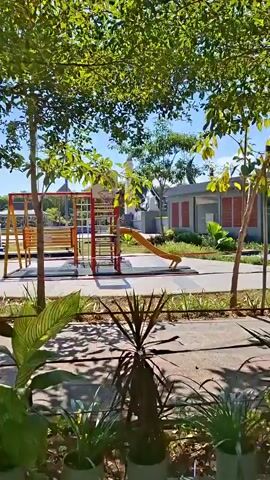The video shows a panning shot of an outdoor children's playground on a sunny day. In the foreground, there are several potted plants. The playground features a colorful climbing structure with red, yellow, and blue components, as well as a yellow slide. A curved yellow bench is visible in the mid-ground. The area is well-maintained with paved walkways and trees providing shade. In the background, there are more trees and some buildings. The overall environment appears peaceful and designed for recreation.