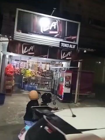 A scooter with two occupants, a driver and a passenger wearing helmets, passes in front of a storefront at night. The storefront appears to be a convenience store named "Toko Alif" with a large illuminated sign. The store is stocked with various items, including what looks like produce and bottled water. Beyond the store, there are several closed shopfronts with green and pink roll-up doors. The street is paved and appears to be dark. A white car is partially visible in the foreground, suggesting the video is taken from inside or from another vehicle. The lighting is dim, indicating it's nighttime or an indoor setting with limited illumination.