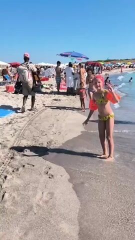 The video captures a lively scene on a sunny beach in Vada, Italy. The main subjects are several people, including children and adults, enjoying a day at the seaside. In the foreground, a young girl in a yellow and green bikini and orange arm floaties performs a cartwheel on the sandy shore, her pink headscarf adding a splash of color. Behind her, another young child in a pink bikini stands watching, and an adult woman in a black bikini is also present.

Further back, on the beach, several other people are visible. A person in a light-colored uniform, possibly a lifeguard or beach attendant, stands near a collection of beach chairs and umbrellas. Other beachgoers are scattered across the sand, some sitting under umbrellas, others walking towards the water. The sea is calm and appears to be a clear, light blue. The sky is a vibrant, cloudless blue, indicative of a warm, sunny day. Beach umbrellas and lounge chairs are set up in rows, suggesting a popular and organized beach area.

The overall mood of the video is cheerful and energetic, highlighting a typical summer day at the beach with families and children engaged in recreational activities. The time of day appears to be mid-morning or early afternoon, given the bright sunlight and the long shadows cast by the people and objects. The location cues, such as the clear blue water and the style of beach setup, are consistent with a Mediterranean beach destination. There is no visible text in the video.