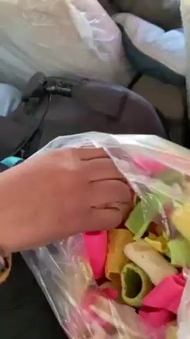 The video shows a person in a car reaching into a plastic bag filled with colorful, tube-shaped snacks. The person takes one out and hands it to another person in the front seat. The person in the front seat, wearing a knit hat and scarf, takes the snack and eats it while looking out the window. The background shows a snowy landscape passing by. The bag of snacks is then shown again, sitting amongst pillows and a bag in the back of the car.