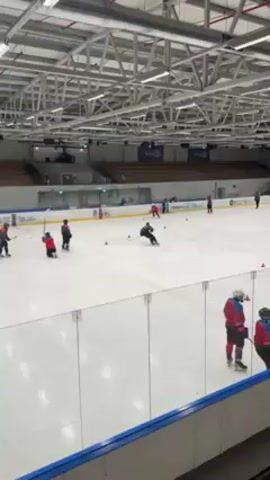 The video shows an indoor ice hockey rink with a group of young people practicing hockey skills. They are wearing hockey gear and using sticks and pucks. Some are skating around cones, practicing maneuvers, while others are engaged in what appears to be a mock game near a net. The rink has white boards around the perimeter and overhead lighting. There is a small, empty spectator area visible in the background. One person is down on the ice for a moment. The overall impression is of a youth hockey practice session.