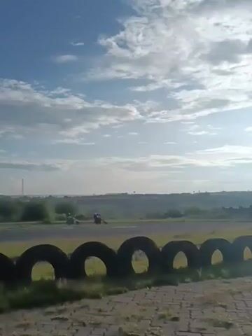 The video shows two motorcyclists speeding along a racetrack, as viewed from behind a tire barrier. The sky above is blue with scattered clouds. The landscape in the background is a mix of fields and some buildings. The camera is relatively stable, providing a clear view of the speeding motorcycles as they navigate a turn on the track.