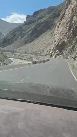 The video shows a car driving on a paved road through a mountainous landscape. A river runs alongside the road. On one side of the road, there are large, blocky concrete barriers. In the distance, snow-capped mountains are visible against a clear, blue sky. A white car passes by in the opposite direction. The shot is taken from inside a car, looking out through the windshield.