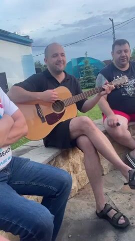 The video shows three men outdoors, with one man in the center playing an acoustic guitar. He is wearing a black polo shirt and black shorts, and his legs are crossed. He is smiling and looking to his left. To his right, another man wearing a black t-shirt and red shorts is seated, looking towards the guitarist. To the left of the guitarist, a third man is visible from the waist down, wearing blue jeans and a white t-shirt with colorful writing. The men are sitting on a low stone wall or ledge in what appears to be a backyard setting. There is a building in the background to the left, and some greenery and a clear structure that might be a greenhouse or awning to the right. The sky is overcast, suggesting late afternoon or evening. The man playing the guitar appears to be in the process of singing or has just finished singing, as indicated by his facial expression and the context of playing an instrument in a social setting. The overall atmosphere seems relaxed and informal.