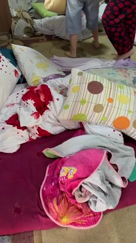 A messy room with a bed covered in blankets and pillows. A woman in a green shirt is sitting on the bed, talking to another woman standing next to her. The second woman is wearing a black shirt and denim shorts and is holding a pillow. The camera then pans to the floor, showing a bare foot and then a woman's legs in a green dress as she walks across the room. The room appears to be a bedroom, with the bed being the central focus. There are several pillows and blankets scattered on the bed, and the floor is covered with a rug. The lighting in the room is dim.