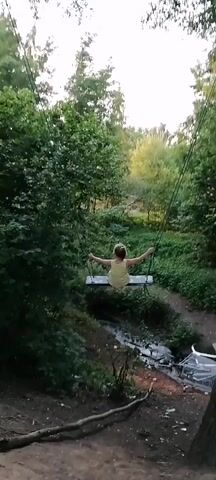 A person is swinging on a swing set in a wooded area. The person is wearing a yellow dress. The swing set is made of rope and wood. The background is a forest with trees and greenery. The lighting suggests it is daytime.