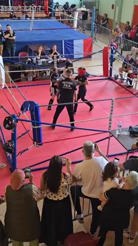 Here's a description of the video:

A high-angle shot shows a kickboxing match in progress in a boxing ring. Two young fighters, wearing protective headgear and gear, are actively engaged in the fight. A referee wearing a black shirt with "REFEREE FKA" is positioned in the center of the ring, closely observing the fighters and ensuring fair play.

The ring is constructed with ropes and padded corners.  The floor of the ring is covered with red mats.  

Around the ring, spectators are seated or standing.  Many are holding up their phones, presumably to take pictures or videos of the match. Several people are visible behind the ring, including a panel of judges.

The setting appears to be a gymnasium or community hall.  A banner with text is visible on the side of the ring.