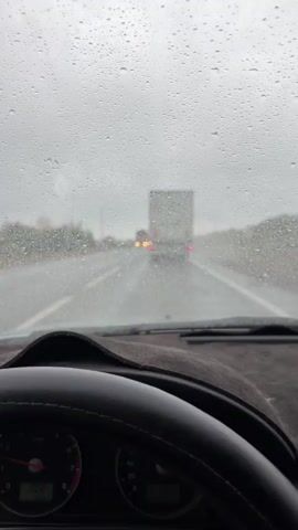 A first-person perspective video shows a driver's view through a rain-streaked windshield on a highway. The road ahead is wet and appears foggy. Two trucks are visible in the distance, one in the left lane and one in the right lane, with their taillights illuminated. There are also some smaller vehicles ahead of the trucks. The driver's steering wheel and dashboard instruments are in the foreground, with a speedometer visible on the right and another gauge on the left. The sky is overcast and gray, indicating rainy weather. The video has a slightly blurry quality due to the rain on the windshield and possibly motion.
