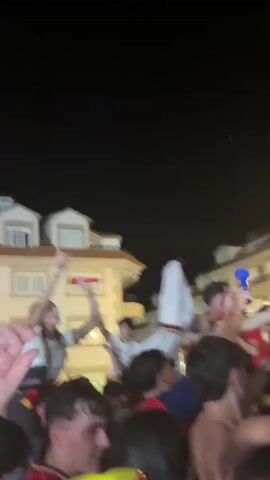 The video shows a crowd of people celebrating at night. There is a large screen in the background displaying an image, possibly a sporting event. A Spanish flag is visible, held by someone in the crowd. People are wearing red and yellow, suggesting they are supporting a team or nation. The atmosphere appears festive and energetic, with people cheering and waving. The buildings in the background are lit up, and the sky is dark. Some individuals are on other people's shoulders, indicating excitement.