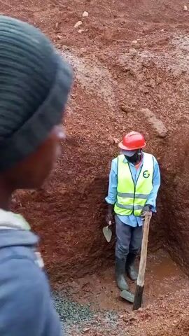 The video shows a man holding a yellow jug standing in a muddy construction site. The man is dark-skinned and wearing a dark blue polo shirt and dark pants. He is also covered in mud. Behind him is a red dirt wall of the construction site. Another construction worker wearing a red hard hat and yellow safety vest stands in the background near the dirt wall. Other workers can be seen in the background. The man with the jug is walking away from the camera. The video appears to be handheld.