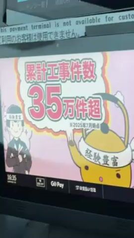 The video shows a close-up of a digital display. The screen displays promotional images and text in Japanese. A message at the top of the screen indicates that the payment terminal is not available for customers in English and Japanese. The promotional content features cartoon characters and text, with phrases like "Speed construction" and "Free 10-year guarantee." The time on the screen is 16:35.