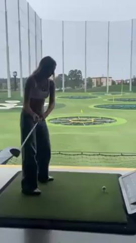 A woman in a white top and jeans is standing on a green golf mat at a Topgolf facility. She swings a golf club, hitting the ball. She then clutches her stomach and laughs. The background shows the typical Topgolf layout with circular targets on an artificial turf field, divided by nets.  There is a screen near the golf mat, presumably displaying game information. It appears to be an overcast day.