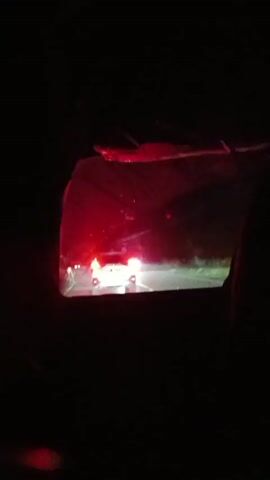 The video is a first-person perspective from inside a vehicle at night, looking out the rear window. In the distance, on a road, a white vehicle with its brake lights illuminated is visible. The scene is dark, with streetlights and the headlights of other vehicles providing illumination. The video appears to be shaky, suggesting movement.