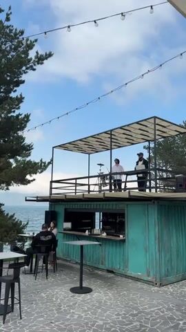 The video shows an outdoor bar area with a DJ setup on an elevated platform. The bar itself is constructed from a teal-colored shipping container. On the elevated platform, two individuals are visible. One person, wearing a white shirt, appears to be operating a drum set. Another person, wearing a black jacket and a beanie, is standing near a laptop, likely acting as a DJ. Below the platform, a bar counter is set up with bottles displayed behind it. Several tables and chairs are arranged in the outdoor seating area. In the background, a body of water, possibly a lake or ocean, is visible, with trees along the shoreline. The sky is blue with some clouds, and string lights are hung overhead.