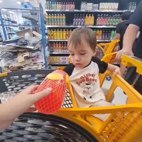 A child sits in a yellow shopping cart. A person’s hand offers a red mesh bag with a yellow object inside to the child. The child reaches out to take it. The shopping cart is in a grocery store aisle. Shelves of bottled drinks are visible in the background. Another person is pushing the shopping cart from behind.