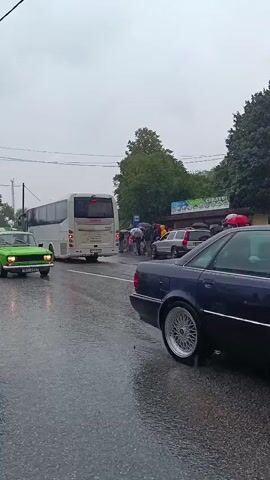 A video captures a street scene on a rainy day. In the foreground, the rear of a dark car is visible on the right side, and the front of a dark car is visible on the left. In the middle ground, a bright green classic car drives past on the wet road. Behind it, a white tour bus is stopped, and several people are gathered around it, some holding umbrellas. Other cars are parked or stopped near the bus. Trees and buildings line the background under a cloudy sky. The road surface is visibly wet and reflective, indicating ongoing rain.