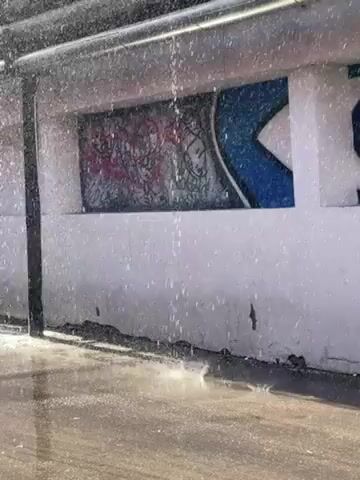 The video shows a close-up view of rainwater falling in front of a white wall adorned with graffiti. A large blue and white eye graphic is visible, along with other less distinct markings. The rain is heavy, obscuring the scene and creating a shimmering effect as it hits the pavement below. A silver pipe or railing is located above, partially visible at the top of the frame. The overall tone is one of urban decay and neglect, highlighted by the damaged wall and the unrelenting rain.