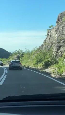The video shows a view from inside a car driving on a paved road. A gray SUV is visible ahead on the right side of the road. The road has white lane markings and is bordered by a guardrail on the right. Steep, rocky hills covered with green vegetation rise on the right side of the road. The sky is clear and blue, and the sun is shining, casting shadows. In the distance, there are rolling hills and mountains. The video appears to be taken on a sunny day in a mountainous, rural area.
