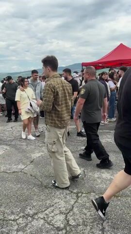 The video shows a large gathering of people outdoors on a paved surface with cracks, possibly an airfield. The sky is cloudy. The crowd consists of both men and women of various ages, dressed in casual clothing. Several individuals wear security uniforms consisting of black shirts and pants. Some people stand in small groups talking, while others walk through the crowd. There are tents and other structures visible in the distance. Some people are wearing high-visibility vests.  The overall impression is that this is a large public event or gathering.