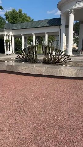 The video shows a fountain in Mariánské Lázně, Czechia. The fountain is made of a dark, metallic material and is shaped like a blooming flower with many petals. Water sprays from the center of the fountain, creating a beautiful display. The fountain is located in front of a colonnade, a covered walkway with columns. In the background, there are trees and a building with a domed roof. The sky is blue and clear, suggesting a sunny day. The ground in the foreground is covered with red gravel. There is no visible text in the video, except for "KOLO" on the building in the background. The overall mood of the video is peaceful and serene.