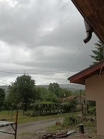 The video captures an outdoor scene during a rainy day in Mihaylovtsi, Bulgaria. The sky is heavily overcast with dark grey clouds, suggesting an ongoing or recent downpour. On the right side of the frame, a portion of a house is visible, featuring a brown-tiled roof and light-colored walls. A dark-colored downspout extends from the roof, actively discharging a steady stream of rainwater.

In the mid-ground and background, the landscape is lush with numerous green trees, some appearing to be part of an orchard or vineyard with trellised plants. A dirt road or path, visibly damp from the rain, runs through the scene. Beyond the trees, other houses are partially visible, and in the far distance, faint outlines of hills or mountains can be seen. Power lines are strung across the upper part of the frame. On the far left, a wooden structure, possibly a table or bench, is partially visible. Near the house on the right, a dark barrel or container and some scattered debris are on the ground. No people are visible, and there is no discernible text. The scene conveys a tranquil, wet atmosphere in a rural or semi-rural setting.