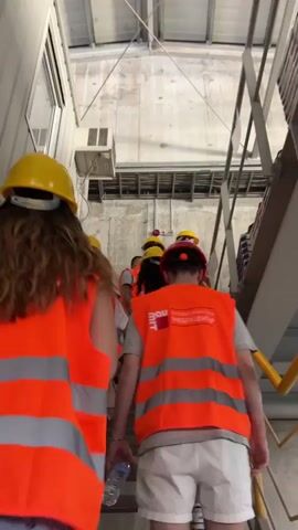 A group of people wearing yellow hard hats and orange high-visibility vests are walking up a metal staircase inside a building. The staircase has yellow railings. The building appears to be an industrial facility, with concrete walls and a metal ceiling. There is a window on the left side of the frame and air conditioning unit mounted on the wall. The people are walking in single file and appear to be on a tour or inspection. The person in the foreground on the left has long brown curly hair and is wearing a blue denim skirt. The person in the foreground on the right is wearing white shorts and a gray t-shirt. There is a bottle of water in their hand. Some of the vests have logos on them.