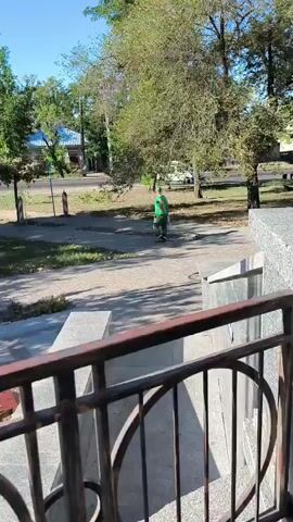 A person in a green shirt and dark shorts is riding a scooter on a paved path. The person is moving away from the camera. The path is surrounded by trees and grass. In the background, there is a building and a road with a few cars. The sky is blue and clear. The video is shot from a slightly elevated position, looking down on the scene.