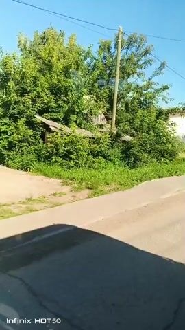 The video shows a view from inside a vehicle moving along a road on a sunny day. A green fence runs along the right side of the road, with houses visible behind it. A pedestrian crossing sign and a road sign indicating "СТРОЙМАРКЕТ" (Construction Market) are also visible. The road surface is asphalt, and there is some gravel and dirt on the side. Lush green trees and vegetation line the left side of the road. The sky is clear and blue. A shadow of the vehicle can be seen on the road surface.