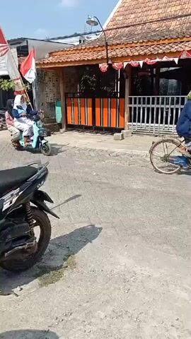 A light blue scooter with two passengers is traveling down a street. The scooter is moving away from the camera and towards a building with a red tiled roof and orange and white striped doors. A flag with red and white stripes is visible on the left side of the screen. A black motorcycle is parked on the left side of the street in the foreground. The street is paved with interlocking bricks. The weather appears to be sunny.