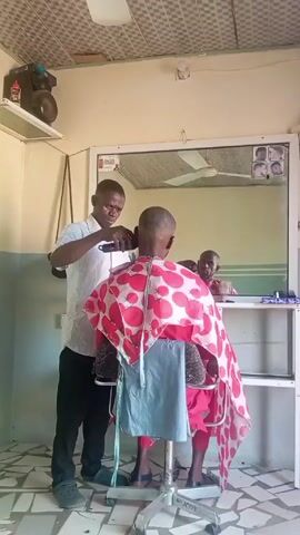 The video shows a barber giving a haircut to a customer inside a barbershop. The barber, a man with short dark hair, wears a light-striped shirt and black pants, along with blue sandals. He is using an electric razor to cut the customer's hair, focusing on the back of the head. The customer is seated in a barber's chair, covered with a red and white polka-dot cape and a light blue towel. The customer's outfit is a red shirt and red pants. 

The barbershop is a small space with cream-colored walls and a patterned ceiling. A large mirror is visible behind the barber and customer, reflecting a second man who is sitting behind a table. The barbershop contains a speaker sitting on a shelf, as well as pictures of haircuts hanging on the frame of the mirror. The floor has linoleum tiles with a green and white pattern.
