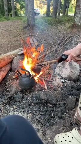 A person's hand is seen holding a black lighter and appears to be igniting a campfire. The campfire has a black kettle placed over it, with flames rising around it. Several rocks surround the campfire, and dry branches and twigs are used as fuel. In the background, a white car is parked on a dirt road, and trees are visible, suggesting an outdoor, wooded environment. The ground is covered with dirt, ash, and scattered debris. Another person's legs are visible in the lower left corner of the frame, wearing dark pants, and another person's feet are visible in the lower right, wearing light-colored clogs. The overall scene depicts a person starting a campfire to heat a kettle outdoors.