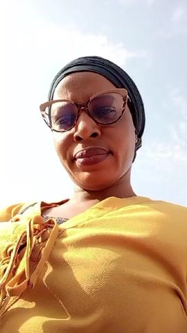 The video features a woman in a mustard yellow shirt, wearing eyeglasses and a black head wrap. She looks directly at the camera from a low angle. The background shows a bright sky with some clouds. There are engine sounds suggesting she might be in or near a moving vehicle.