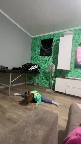 A young person with blonde hair wearing a green sweatshirt and blue pants is performing a split on a wooden floor. They are lying face down with their legs extended to the sides. The environment appears to be a living room, with a couch in the foreground, a massage table to the left, and a wall with green textured wallpaper behind the person. A fan and a cabinet are also visible against the green wall, and a television is on to the right.