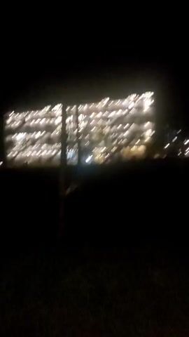 The camera focuses on a large building brightly lit at night. The building has multiple floors and windows, indicating a residential or commercial structure. The foreground is dark, possibly a field or open space, with some blurred silhouettes of what could be trees or vegetation. A hand briefly enters the frame with a peace sign. The scene is static, with no evident movement other than the fleeting hand gesture.
